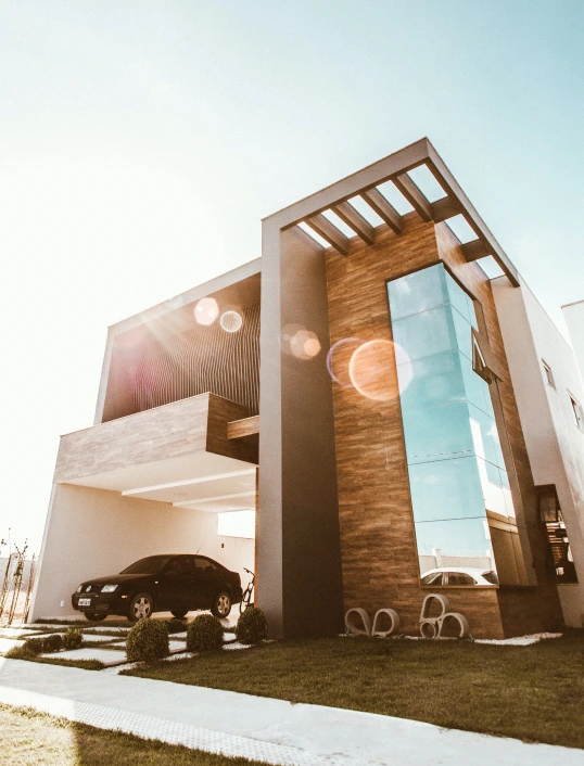 Modern two-story house with large glass windows and wooden accents, featuring a sleek black car parked in front. Sunlight creates a warm glow.
